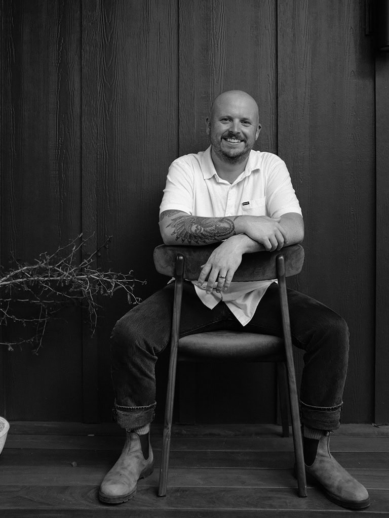 Sam McNees, a man with a shaved head and a beard sits casually on a wooden chair, smiling warmly. He is wearing a white shirt and boots, against a wooden wall background.