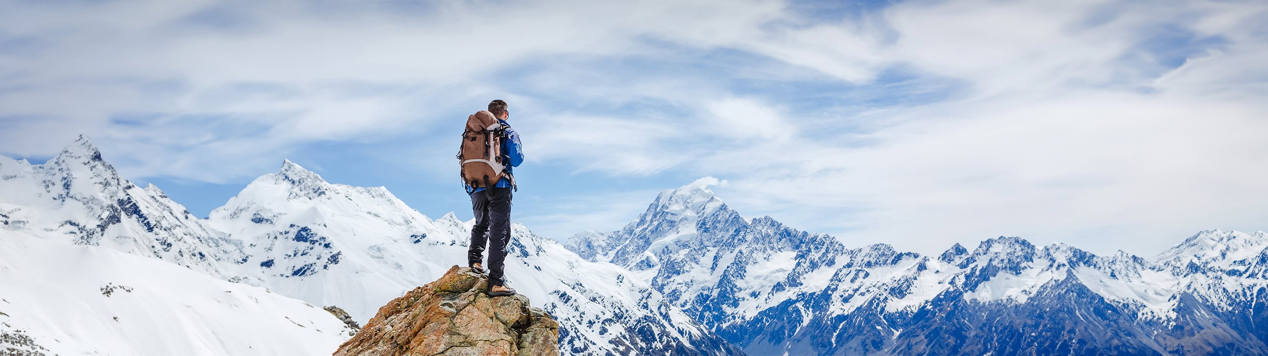 A hiker in a blue jacket and brown backpack stands on a rocky peak, overlooking snow-covered mountains under a partly cloudy sky, capturing a moment of adventure and awe in nature.