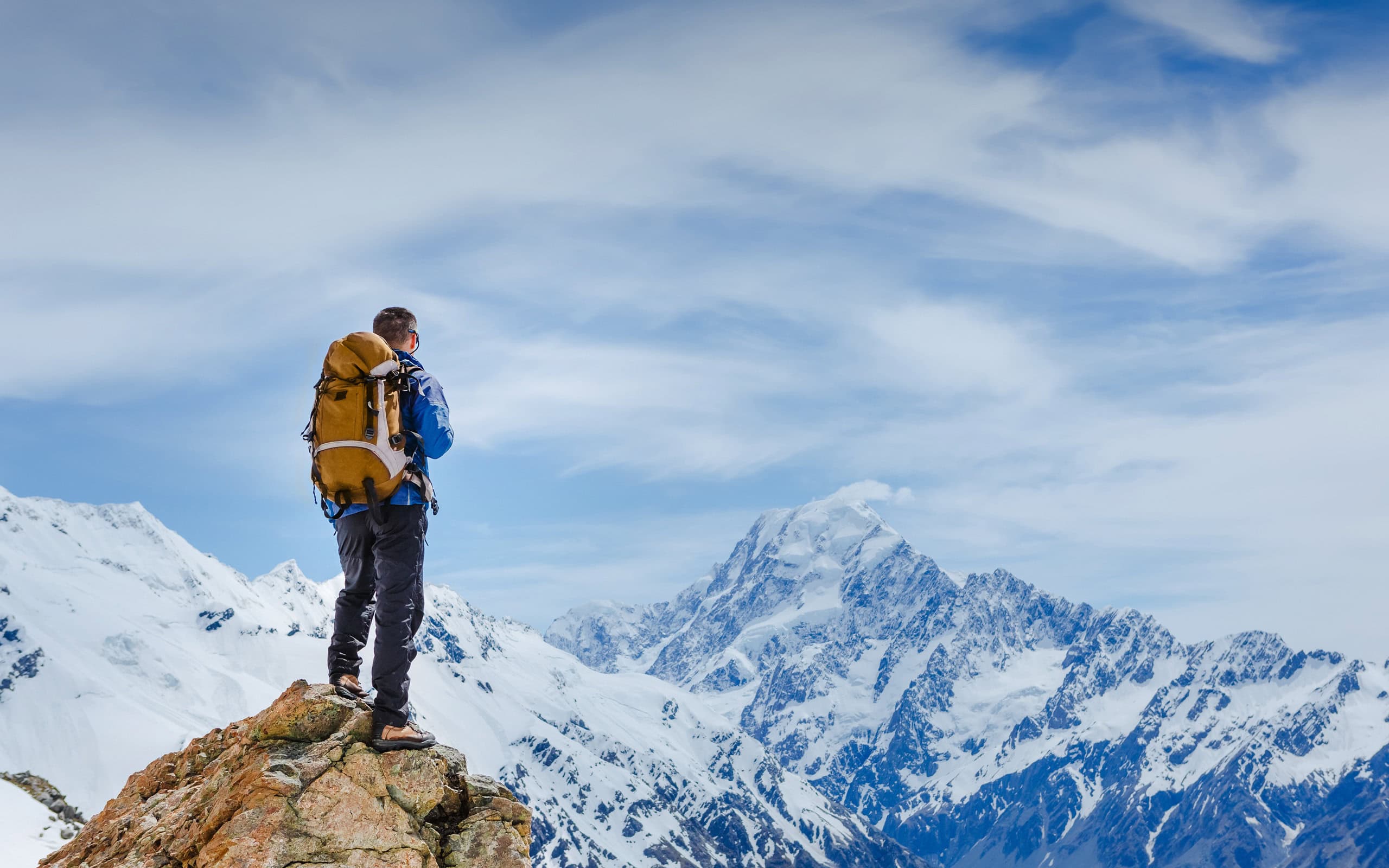 A hiker in a blue jacket and brown backpack stands on a rocky peak, overlooking snow-covered mountains under a partly cloudy sky, capturing a moment of adventure and awe in nature.